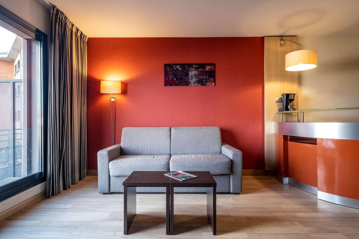 A modern hotel room with a grey sofa, a small table with a magazine, a floor lamp, and a coffee machine.