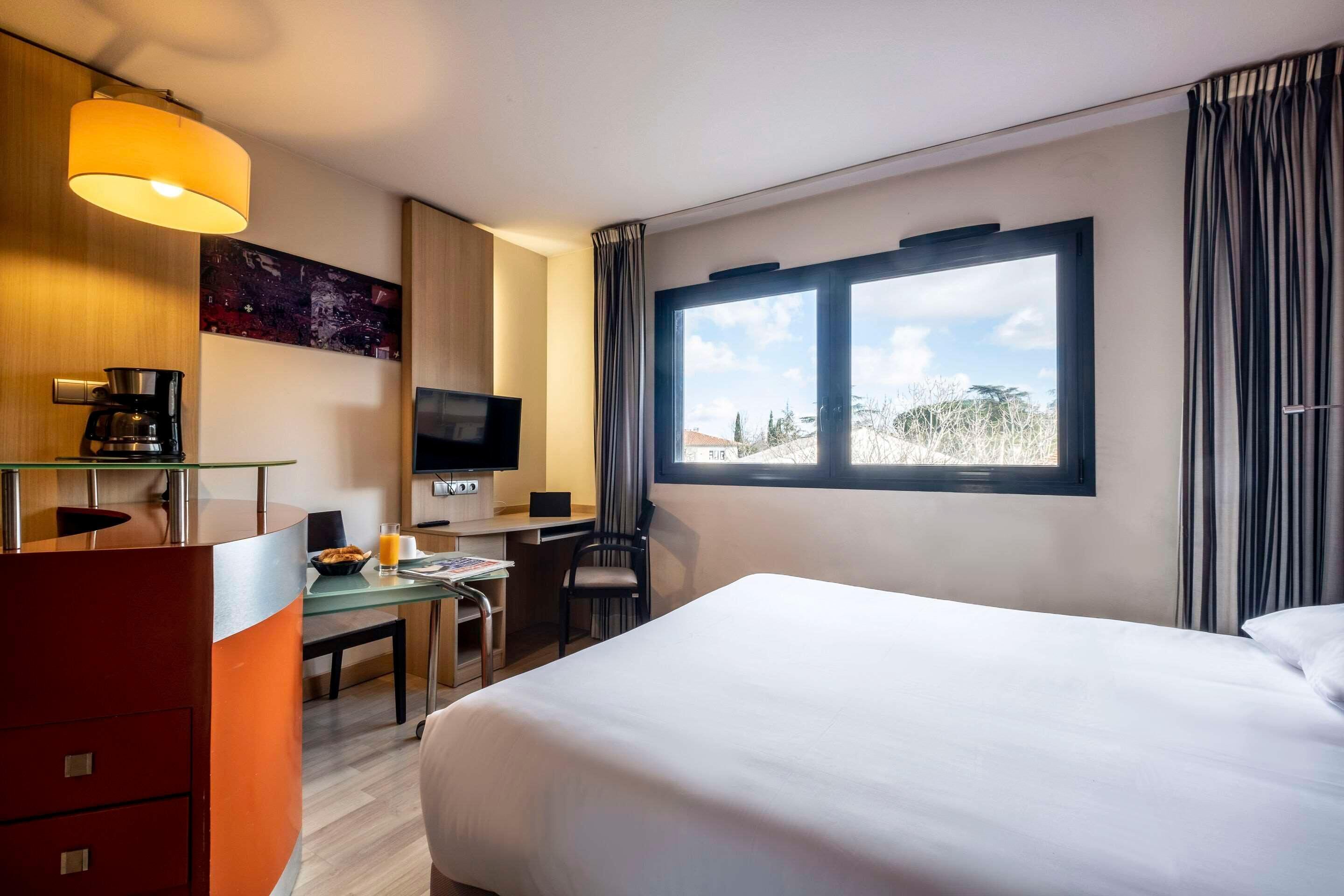 A hotel room with a bed, a desk with a computer, a TV, a coffee machine, and a window with curtains.