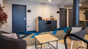 A modern hotel room with a sofa, armchair, and a small table. There is a kitchenette with a coffee machine and a fruit basket. The room has a blue and gray carpet and a large pillar.