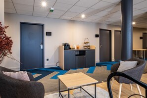 Una habitación de hotel moderna con sofá, sillón y una pequeña mesa. Cuenta con una kitchenette con cafetera y una cesta de fruta. La habitación tiene una alfombra azul y gris, y un pilar grande.