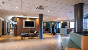 A modern lobby with a wooden reception desk, a seating area with a sofa and a table, and a large TV screen.