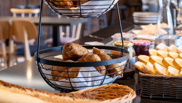 A buffet setup with a three-tiered metal rack holding pastries, a wicker basket with bread, and a variety of other food items on a table.