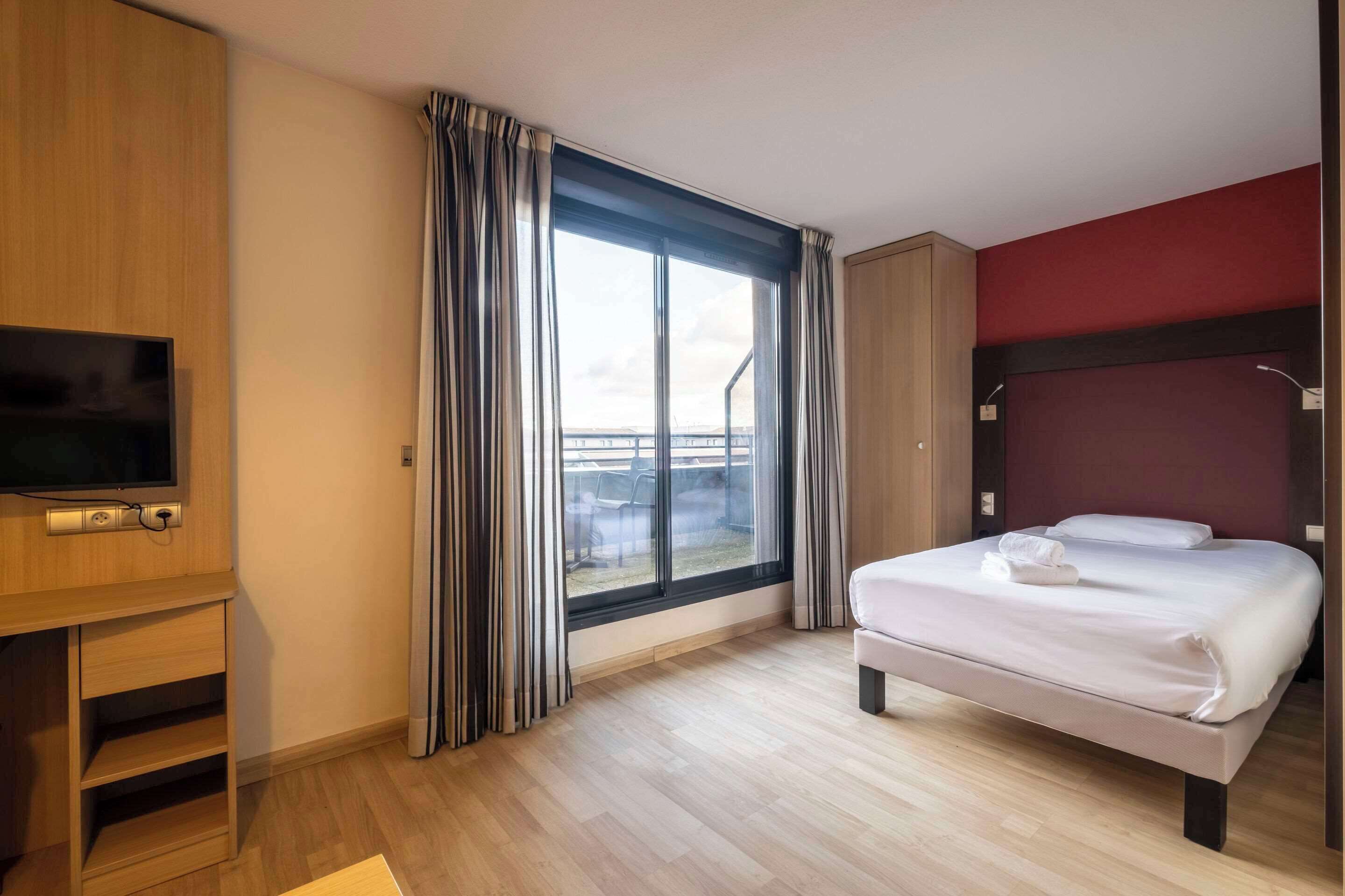 Habitación de hotel con cama, televisor, ventana con cortinas y vistas al exterior.