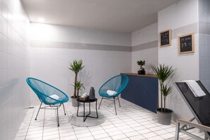 Un baño moderno con azulejos blancos, dos sillas azules, una mesita con una planta y una tetera, y un perchero para toallas.