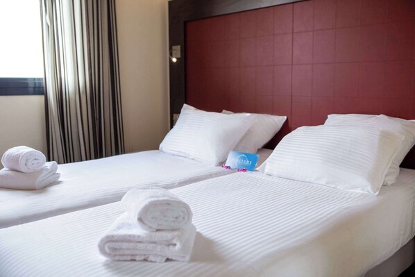 Habitación de hotel con dos camas, sábanas blancas, toallas dobladas con cuidado y un cabecero rojo.