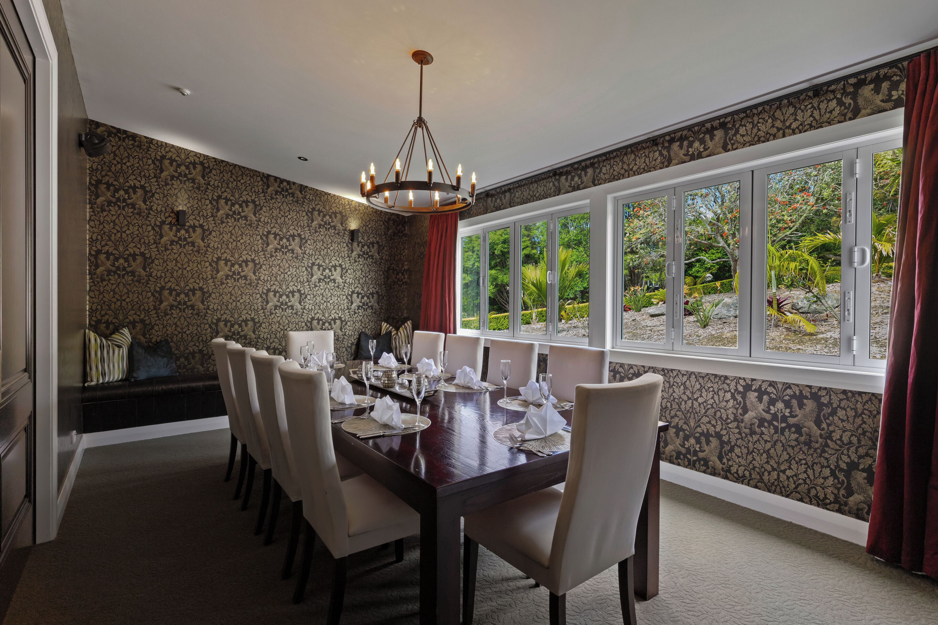 Een eetzaal met een grote tafel gedekt voor een maaltijd, een kroonluchter en uitzicht op groen via grote ramen.