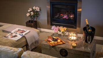 A cozy living room with a fireplace, a magazine, a wooden tray with snacks, a candle, and a bottle of champagne.
