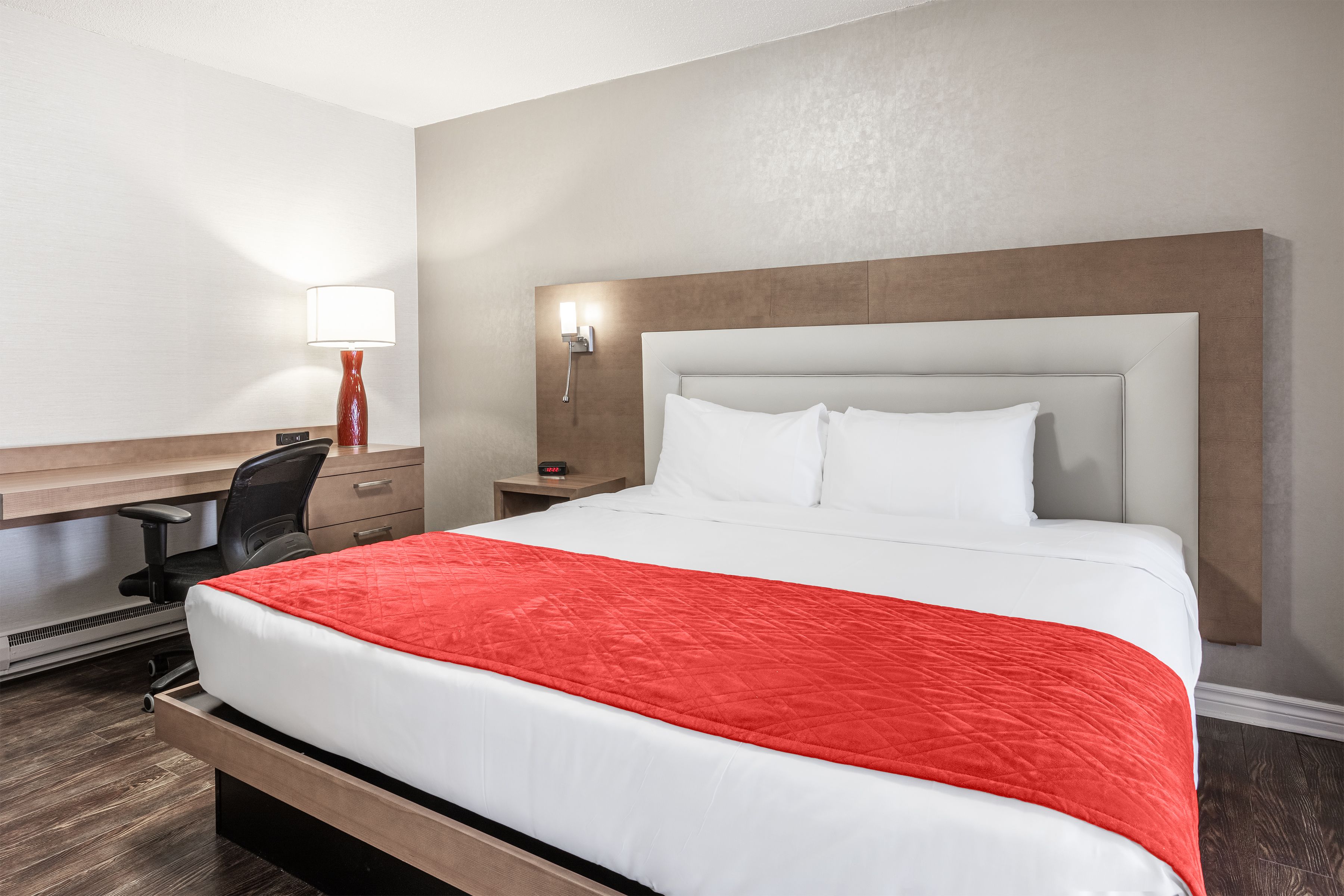Une chambre d’hôtel équipée d’un lit, d’un bureau, d’une chaise, d’une table de chevet, d’une lampe et d’une horloge murale.