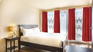 Habitación de hotel con cama, mesita de noche con lámpara, ventana con cortinas rojas y vistas al exterior.