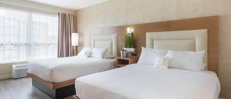 Habitación de hotel con dos camas, un ventanal con cortinas, una mesita de noche con lámpara y una planta en la pared.