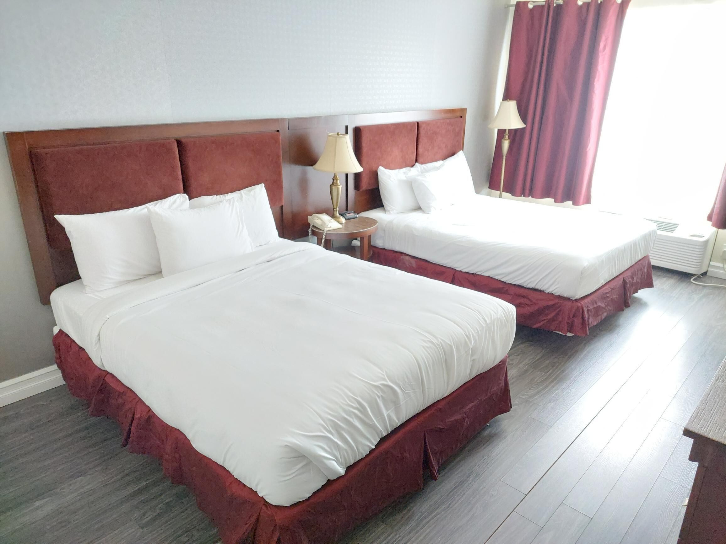 Habitación de hotel con dos camas, cada una con cabecero de madera y ropa de cama blanca. Se observa una mesita de noche con lámpara y teléfono.