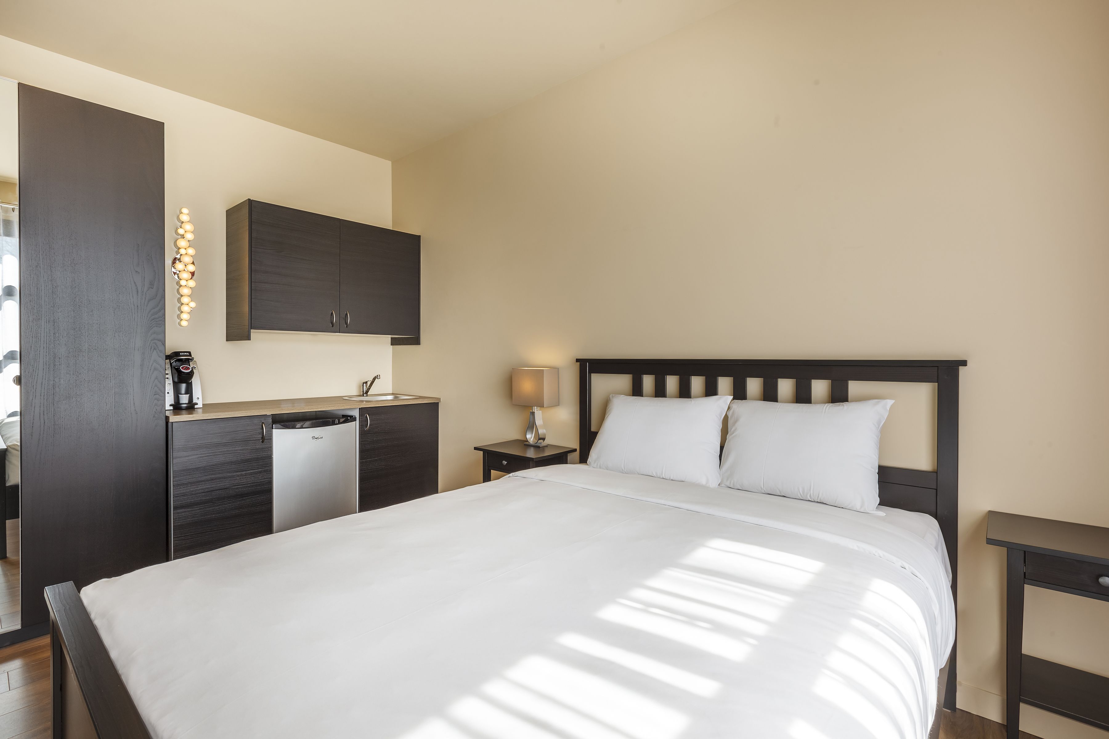 A bedroom with a bed, a nightstand, a lamp, a wardrobe, and a built-in desk with a coffee maker.