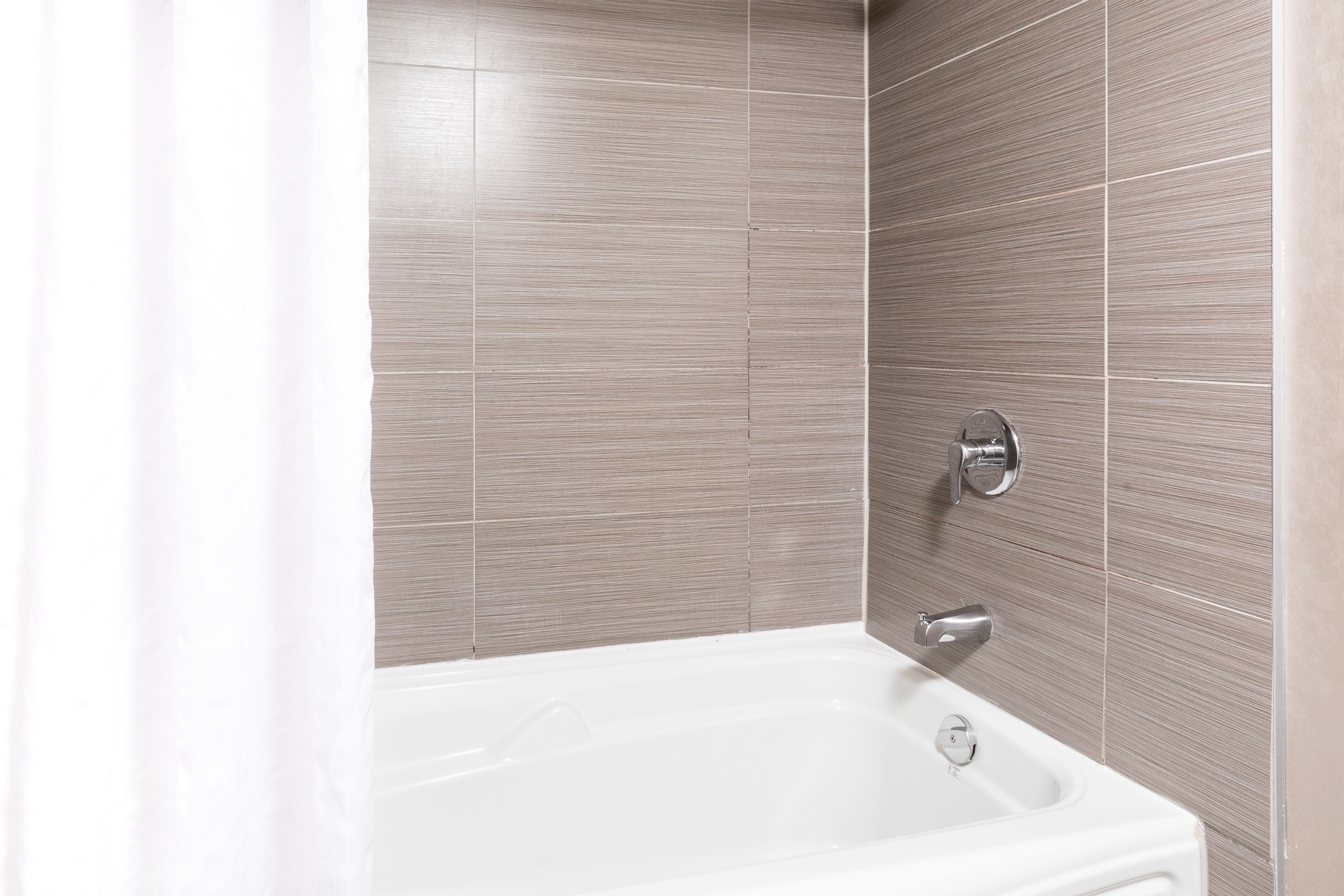 Un baño con una bañera blanca, un grifo de cromo y paredes cubiertas de baldosas beige.