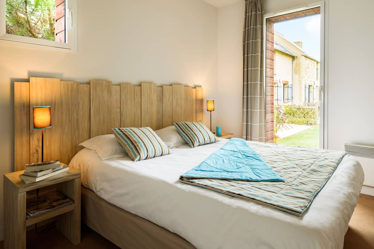A neatly made bed with striped pillows and a blue blanket, wooden headboard, bedside lamps, and a view of a building through a window.