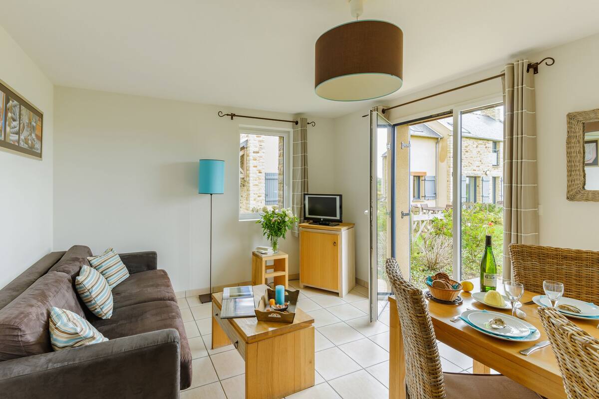 A living room with a sofa, coffee table, TV, and dining area with wicker chairs and a table set for two.