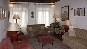 A living room with striped sofas, armchairs, and a red ottoman. There are two lamps on the floor, a potted plant, and framed pictures on the wall. - Hotel Riviera (Évora)