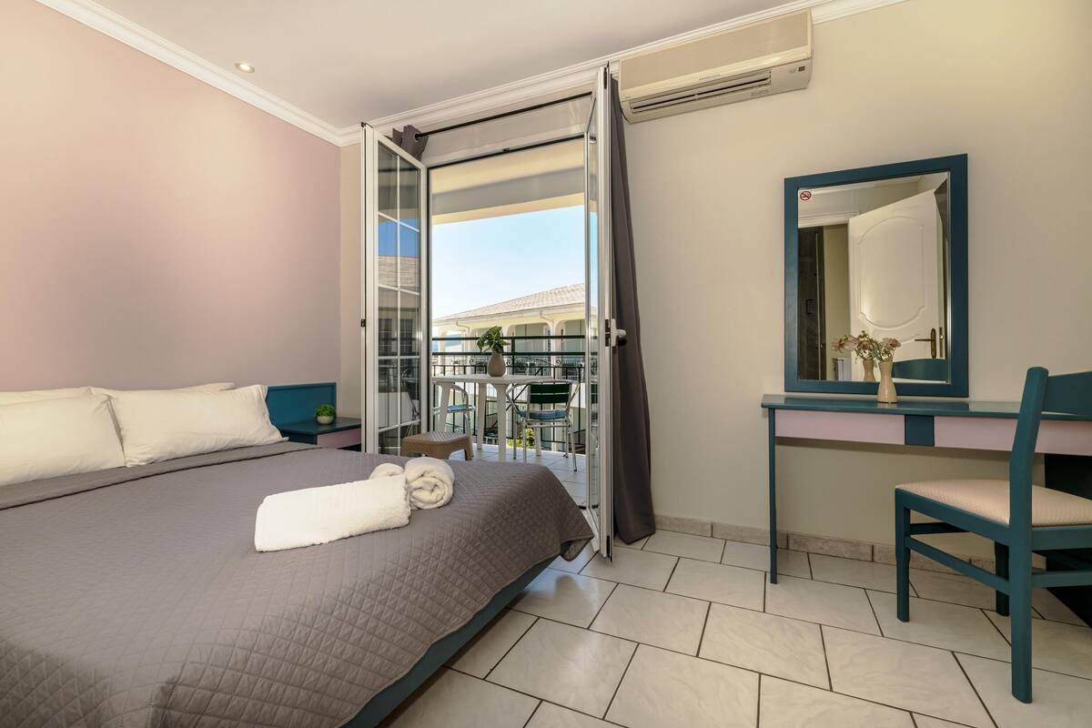 Ein Hotelzimmer mit Bett, Schreibtisch, Stuhl, Spiegel und einem Balkon mit Blick auf ein Gebäude.