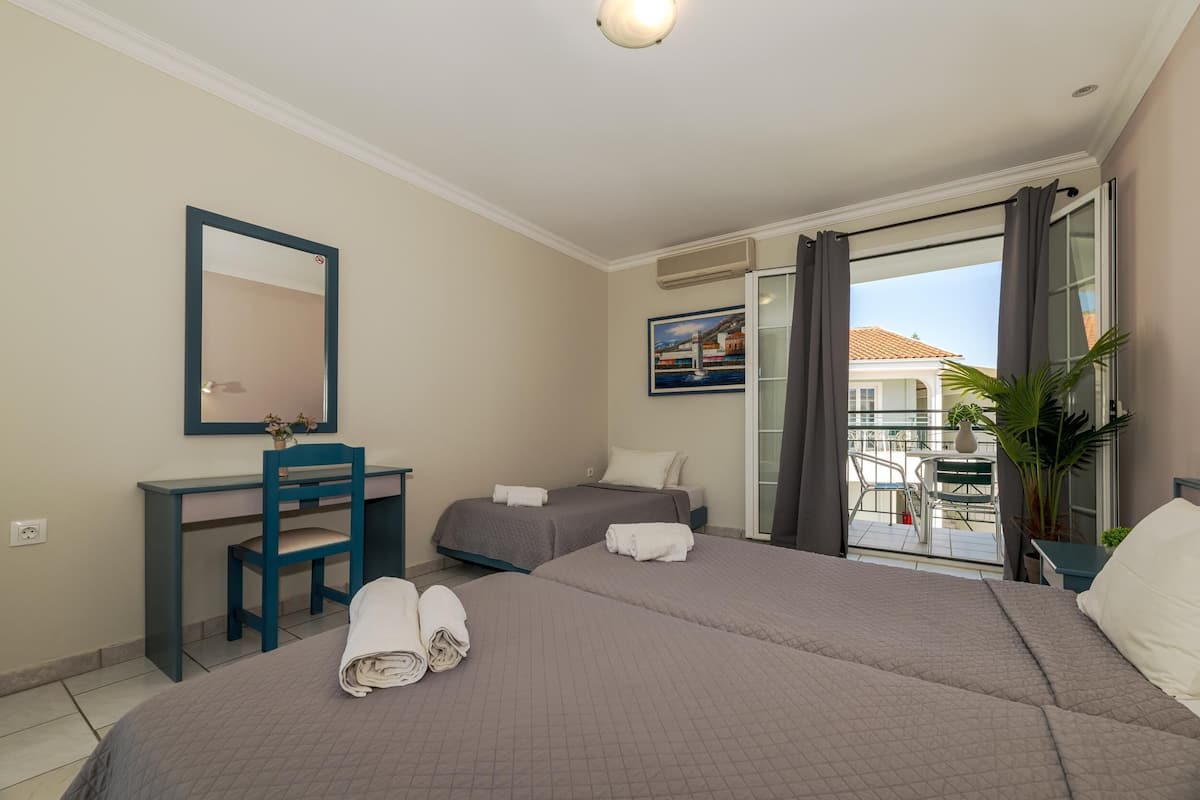 Ein Hotelzimmer mit zwei Betten, einem Schreibtisch, einem Sessel und einem großen Spiegel, sowie einem Balkon mit Blick auf Gebäude.