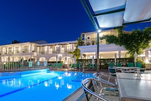 A swimming pool with lounge chairs and umbrellas, a covered seating area, and a building with multiple balconies and windows.
