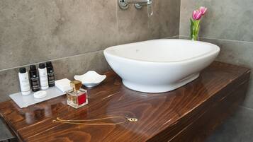 Un baño con un lavamanos grande de color blanco, un mueble de tocador de madera y artículos de aseo, incluyendo una botella de perfume.