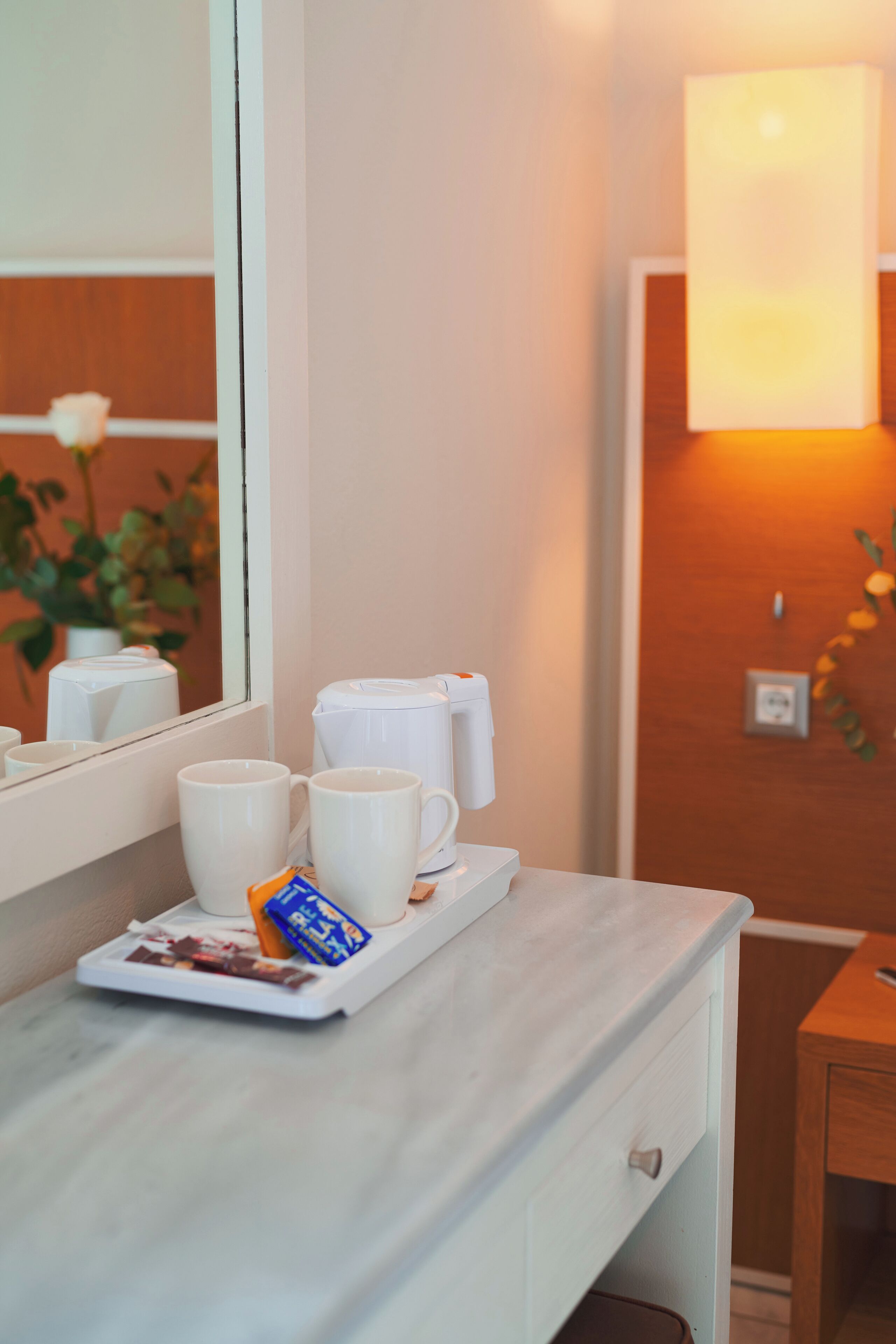 A hotel room with a white marble counter, a tray with two white mugs and a chocolate bar, a white coffee maker, and a wall-mounted light fixture.