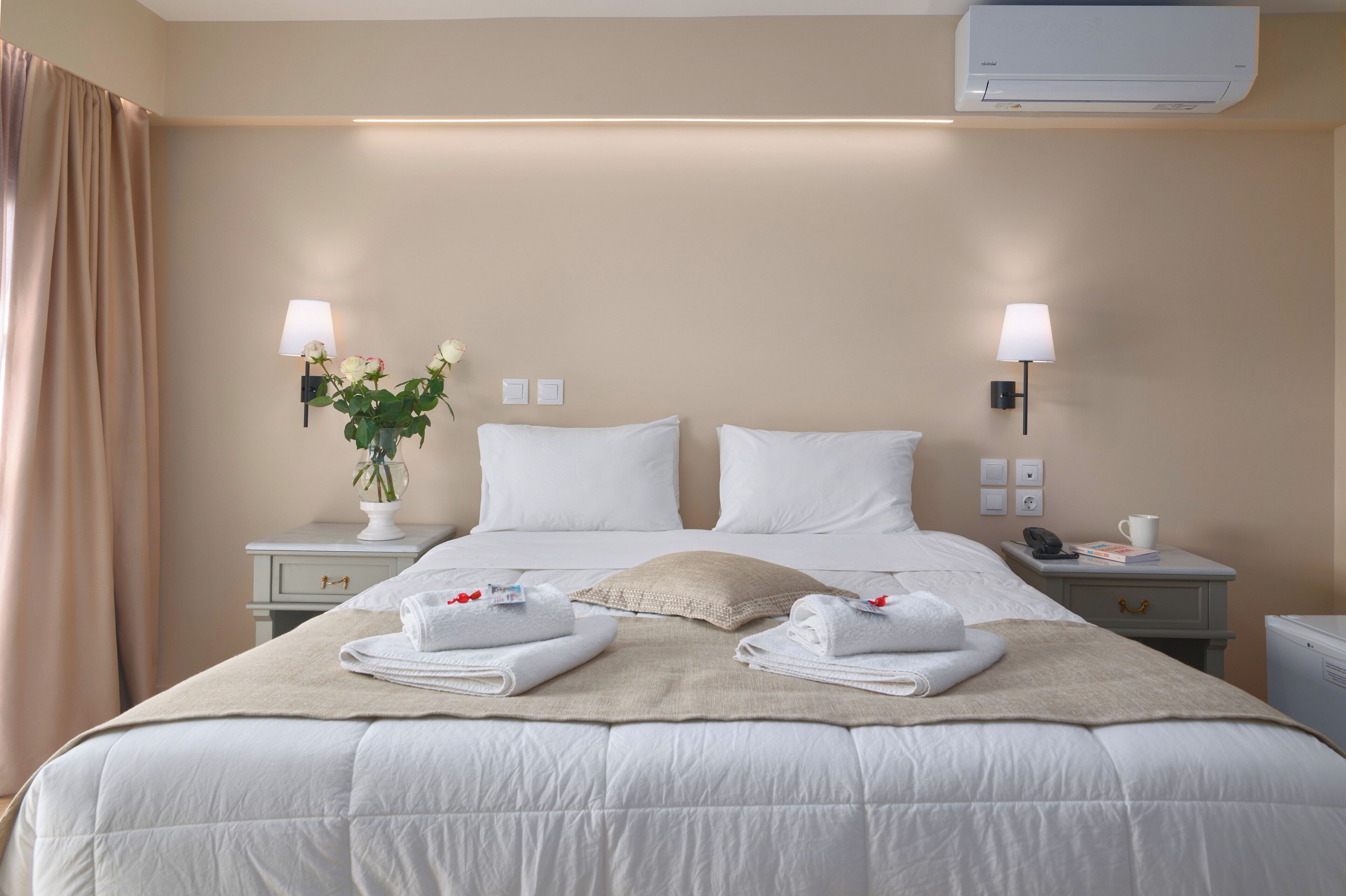 A hotel room with a large bed, two bedside tables, a nightstand, a wall-mounted air conditioner, and a window with curtains.