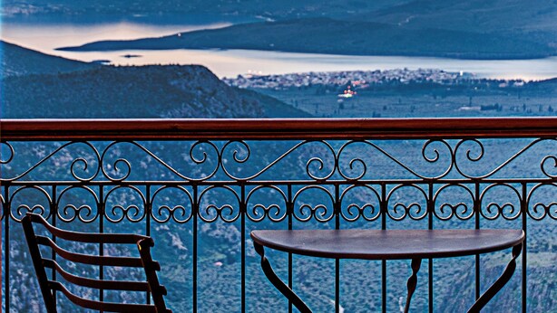 Un balcon avec une table et des chaises, d’où l’on peut admirer un paysage montagneux avec un lac et une ville au loin.