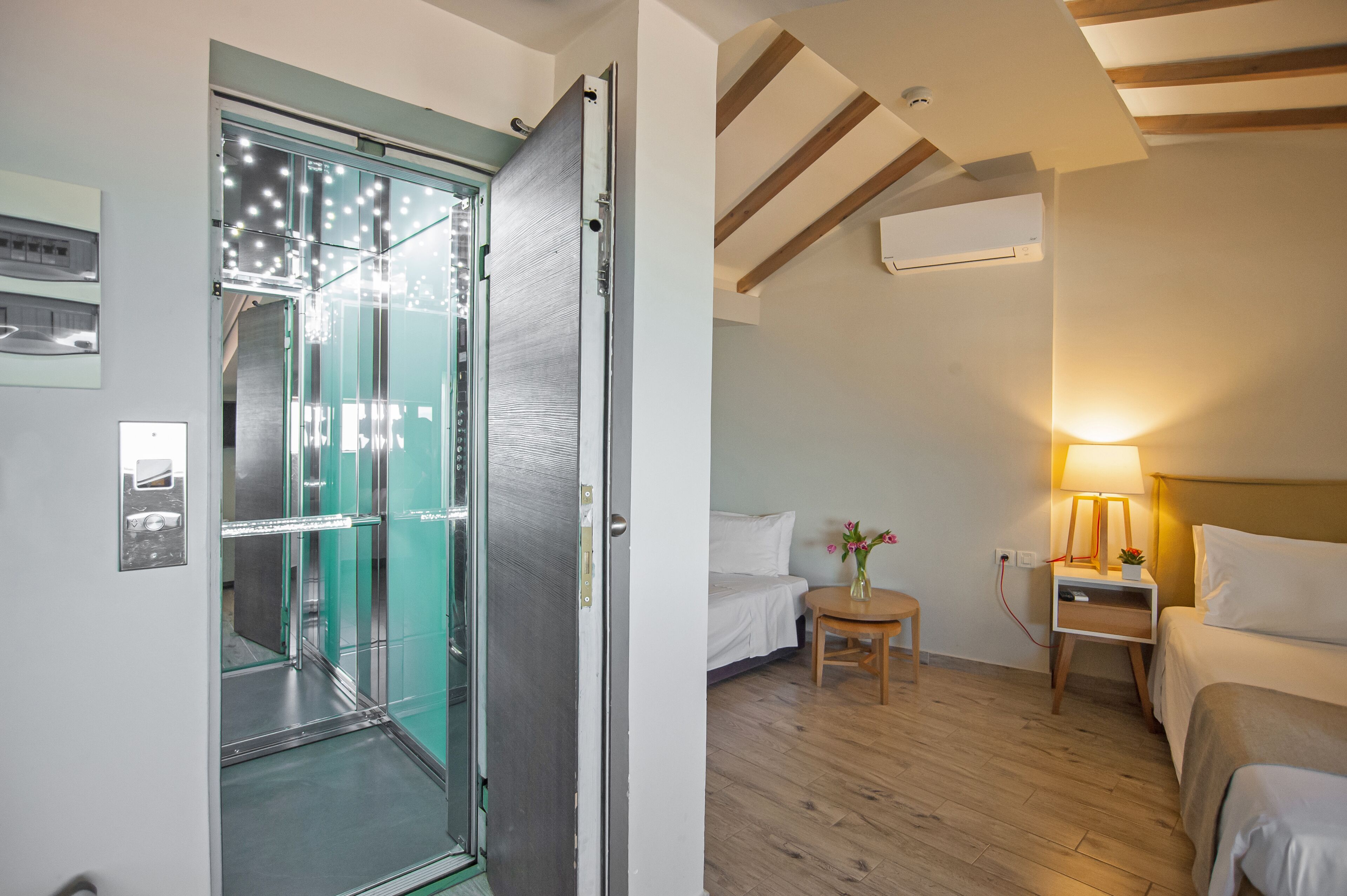Habitación de hotel con ascensor de vidrio, una cama, mesita de noche con lámpara y una pequeña mesa con flores.