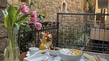 Ein Balkon mit einer Steinmauer, einem Metallgeländer und einem Tisch, gedeckt mit Frühstücksgegenständen, darunter ein Glas Orangensaft, eine Schale mit Obst und ein Teller mit Eiern.