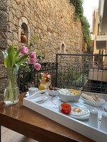 Un balcón con pared de piedra, barandilla metálica y una mesa dispuesta para el desayuno, con un vaso de jugo de naranja, un bol de fruta y un plato de huevos.