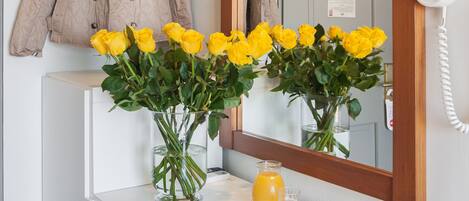Une salle de bain avec un meuble-lavabo blanc, un miroir, un manteau accroché, un vase de fleurs jaunes, un verre de jus d’orange et un magazine.