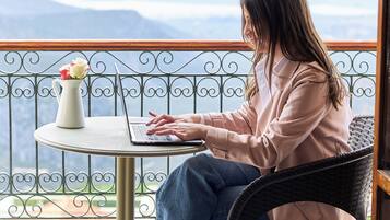 En person sidder ved et bord med en bærbar computer, iført en let jakke og jeans, på en balkon med udsigt til bjerge.