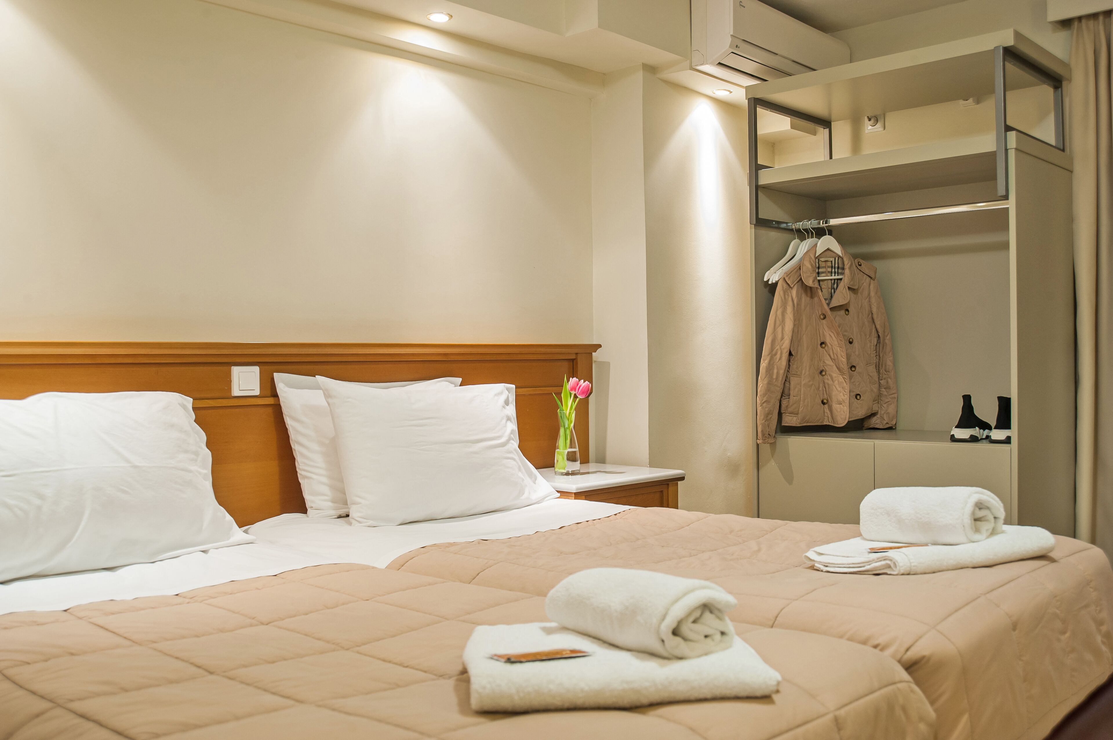 A hotel room with two beds, a wooden headboard, a nightstand with a vase of flowers, and a mirror with a hanging coat.