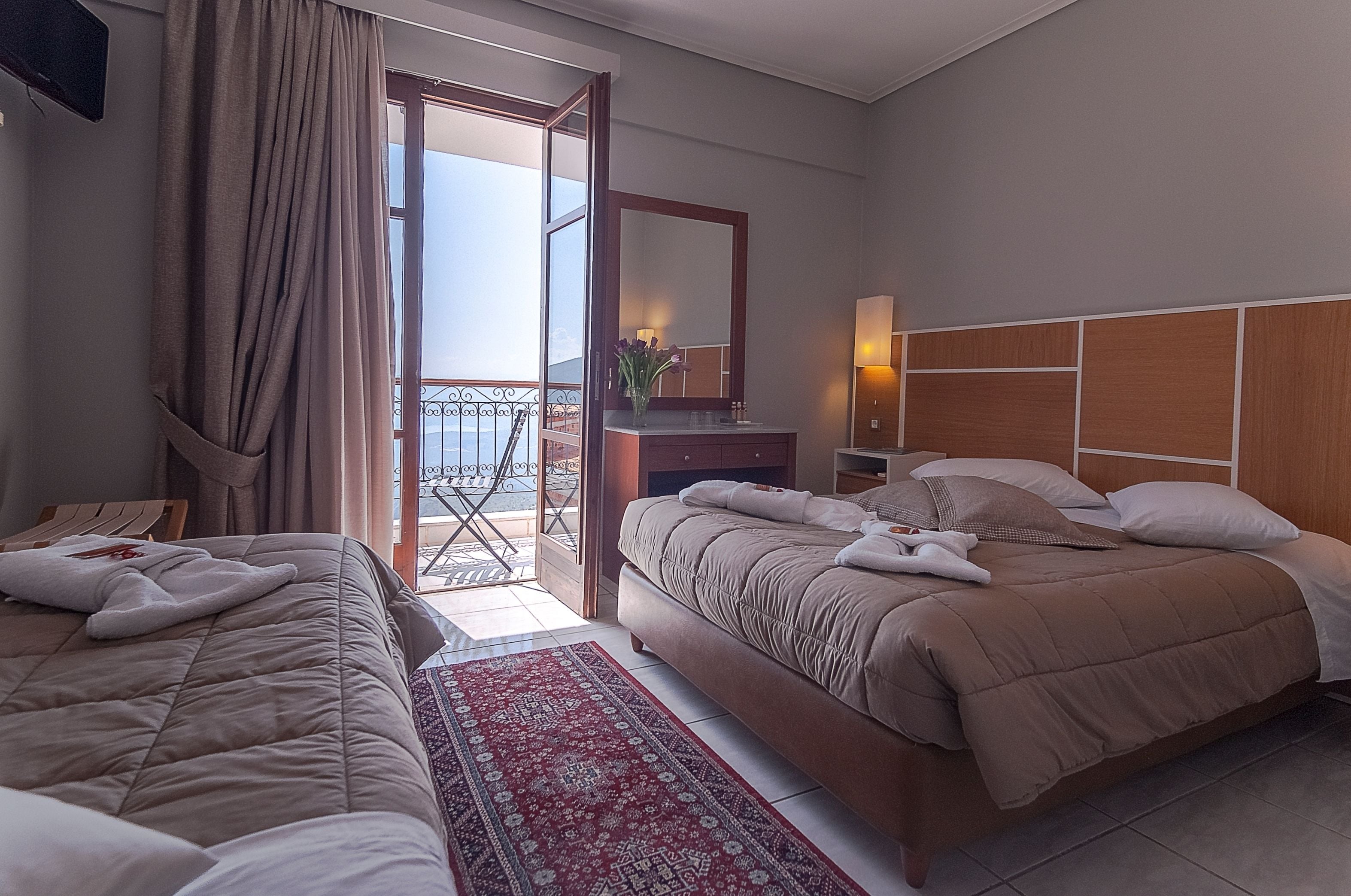 Habitación de hotel con dos camas, un tapete rojo estampado, un balcón con vistas y un cabecero de madera.