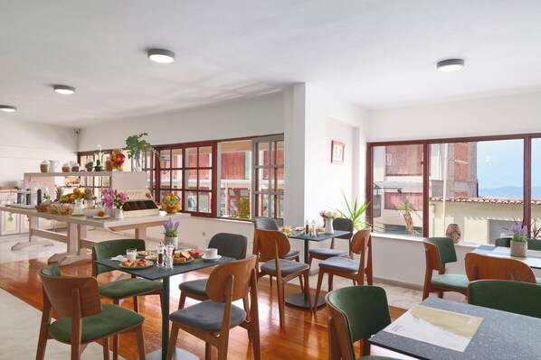 A spacious dining area with multiple tables and chairs, a kitchen area with various items on display, and large windows offering views of the outside. - Fedriades Delphi Hotel (Delphi)