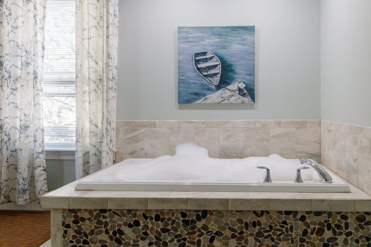 A bathroom with a mosaic-tiled sink area, a painting of a boat on a dock, and a window with floral curtains.