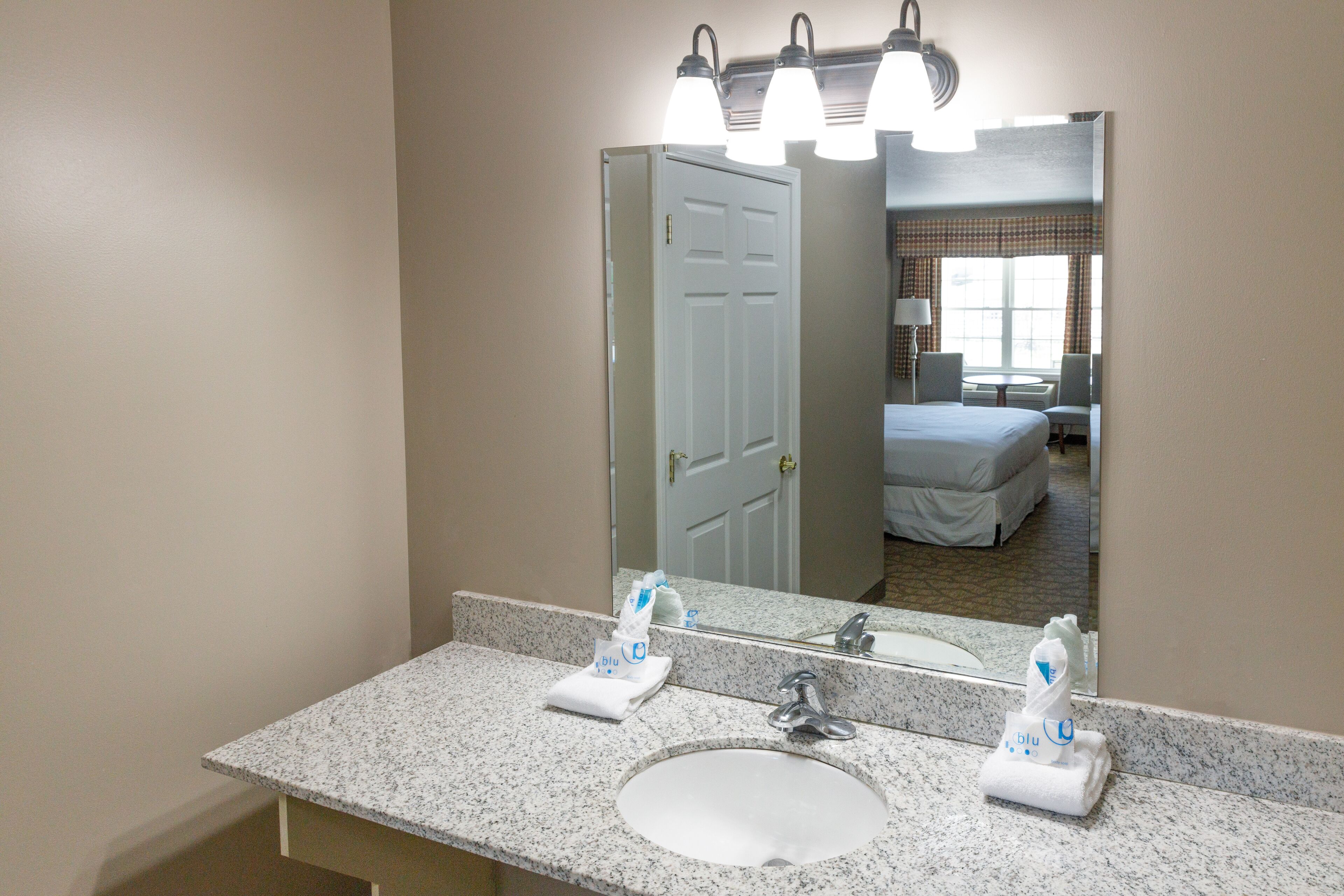 A hotel bathroom with a granite countertop, a sink, a mirror, and a door leading to a bedroom.