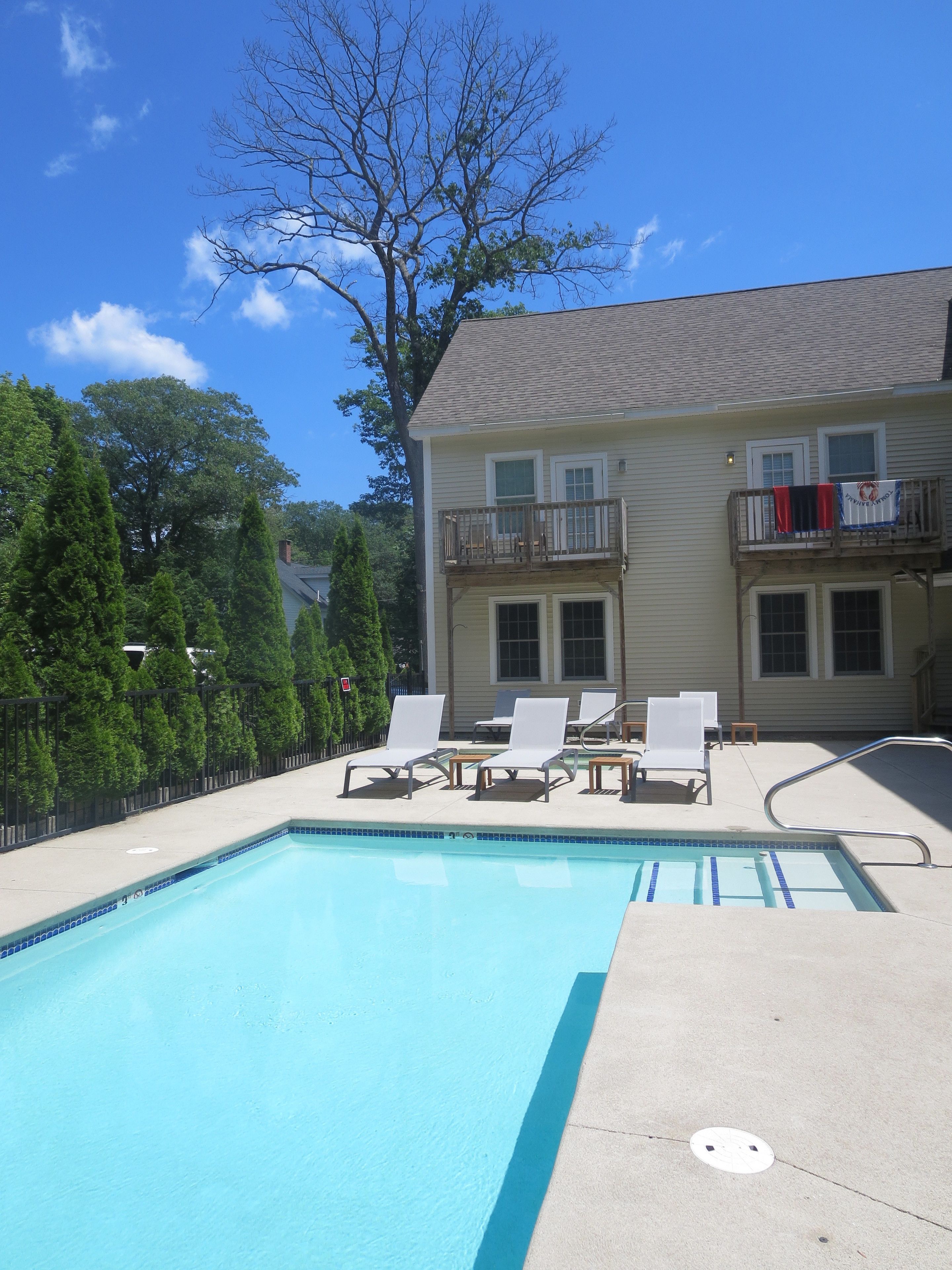 A swimming pool with lounge chairs, a two-story house with a balcony, and a large tree in the background.