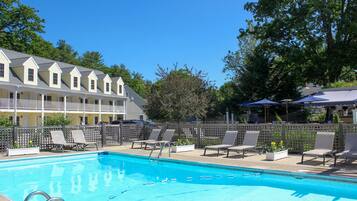 A swimming pool with lounge chairs and umbrellas, surrounded by a wooden fence and a building with multiple windows.