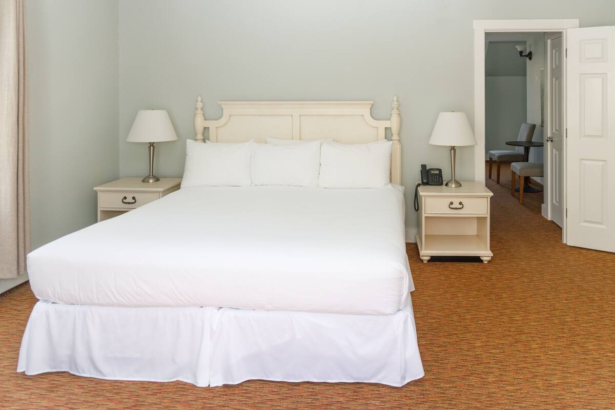 A neatly arranged bedroom with a large bed, two bedside tables with lamps, a chair, and a door leading to another room.
