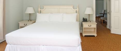 Un dormitorio ordenado con una cama grande, dos mesitas de noche con lámparas, una silla y una puerta que da a otra habitación.