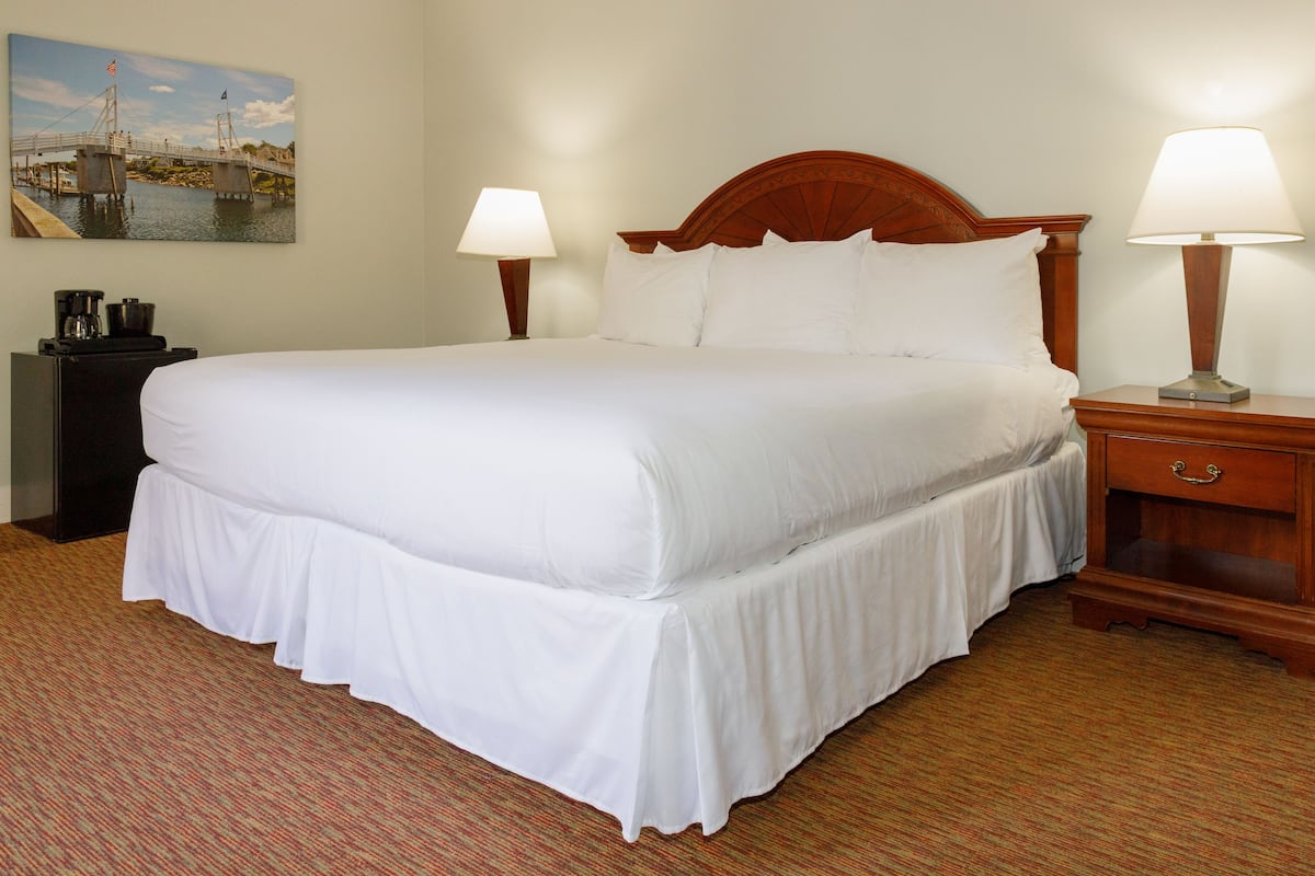 A neatly made bed with white linens, a wooden headboard, two bedside lamps, a nightstand with a drawer, and a painting of a bridge on the wall.