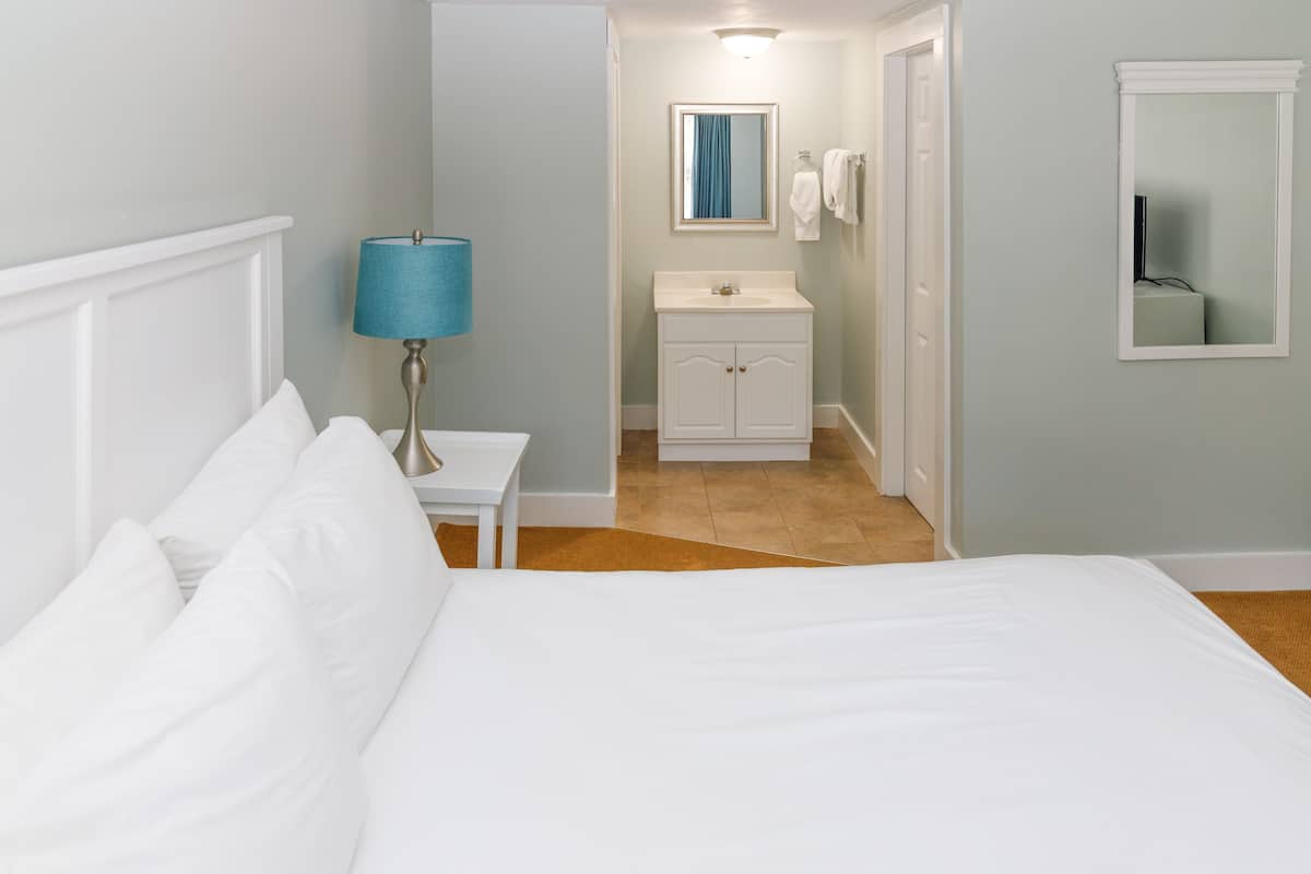 A bedroom with a bed, a nightstand, a lamp, a bathroom with a sink, and a mirror.