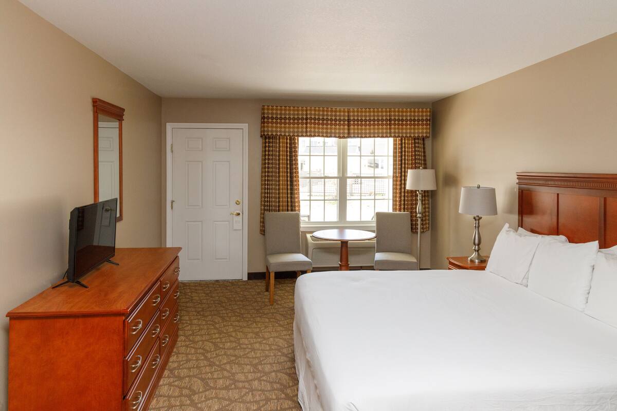 A hotel room with a bed, a dresser, a television, a desk with a chair, a lamp, and a window with curtains.