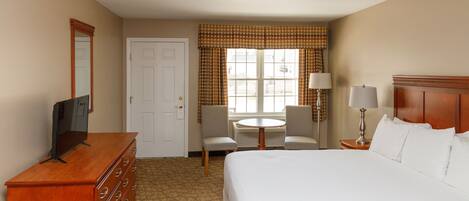 A hotel room with a bed, a dresser, a television, a desk with a chair, a lamp, and a window with curtains.