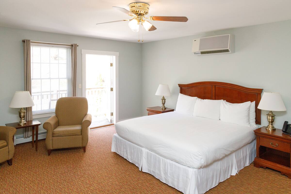 A hotel room with a large bed, two bedside tables with lamps, a chair, a ceiling fan, and a window with curtains.