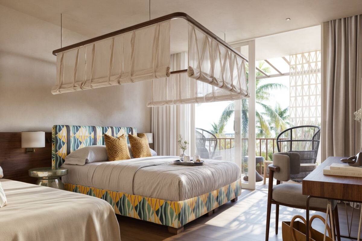 Hotelkamer met een groot bed, een kleiner bed, een bureau, een stoel en een balkon met uitzicht op palmbomen.