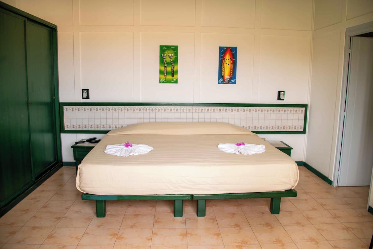 Een tweepersoonsbed met een groen hoofdeinde en voeteinde, twee handdoeken, een nachtkastje in het groen, een telefoon en twee ingelijste kunstwerken aan de muur.