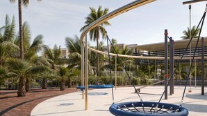 Un parque infantil con columpios y una zona de estar circular, rodeado de palmeras y un edificio al fondo.