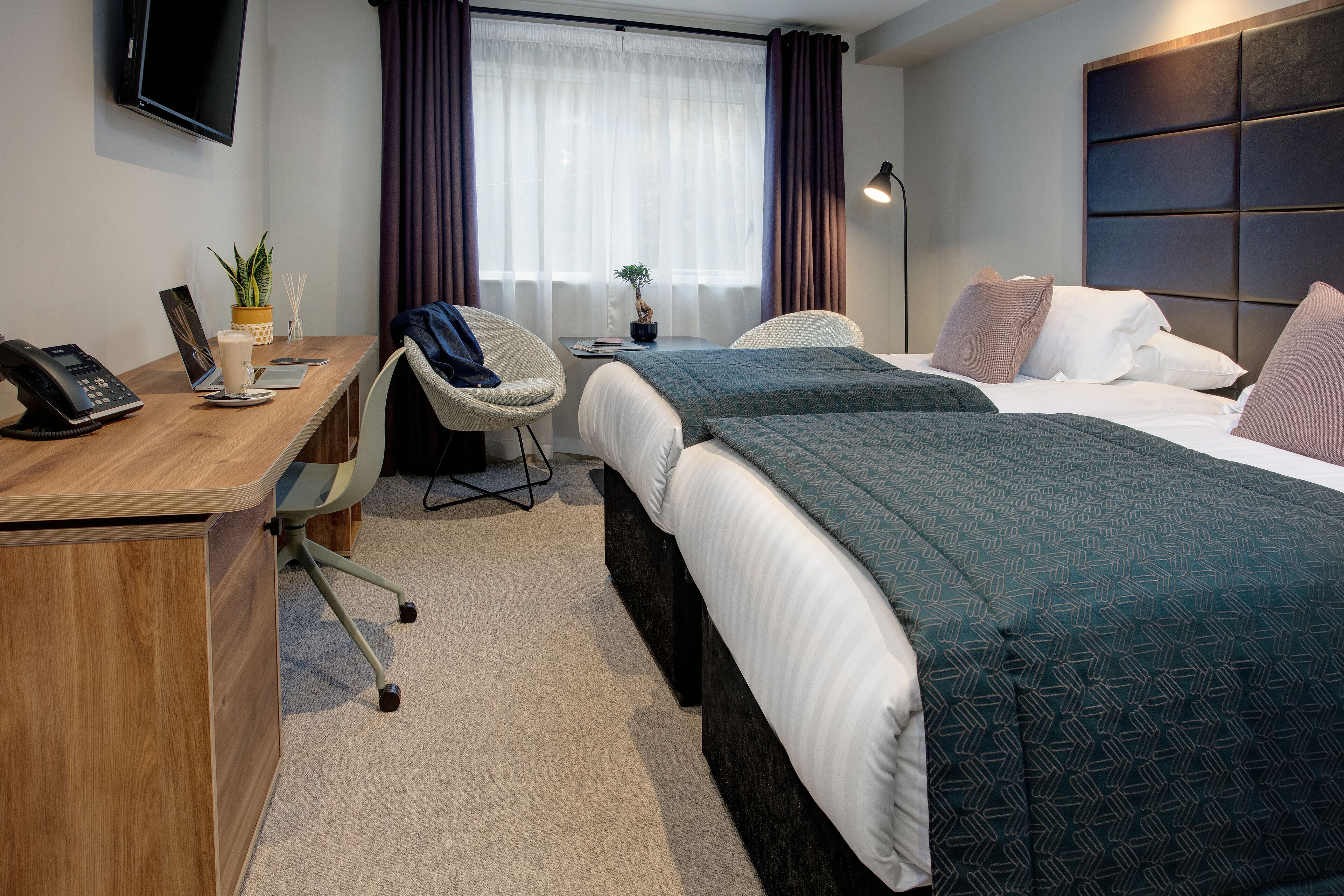 A hotel room with a wooden desk, two beds, a chair, a TV, and a window with curtains.